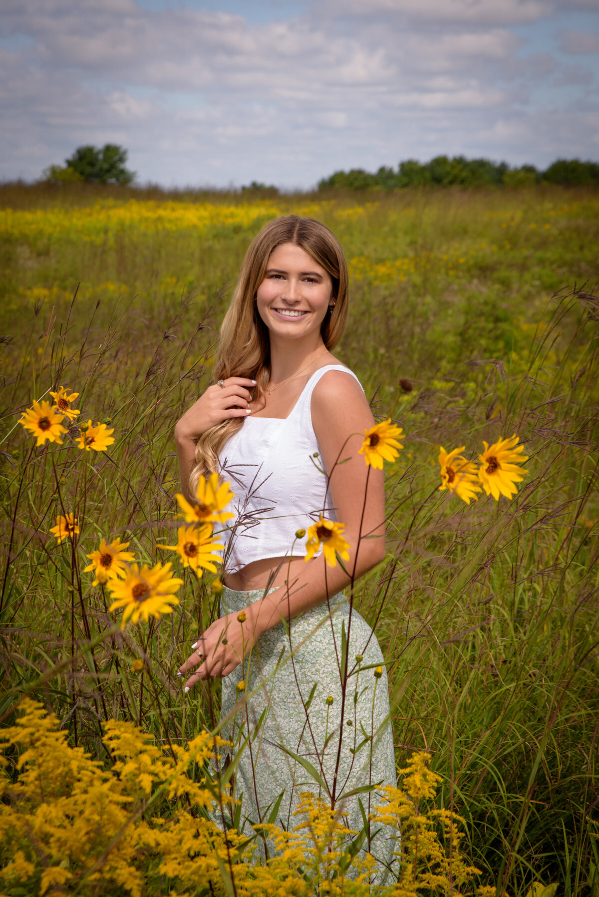 De Pere High School senior girl wearing long green skirt and white shirt standing in long grassy field and wildflowers at Fonferek Glen County Park in Green Bay, Wisconsin.