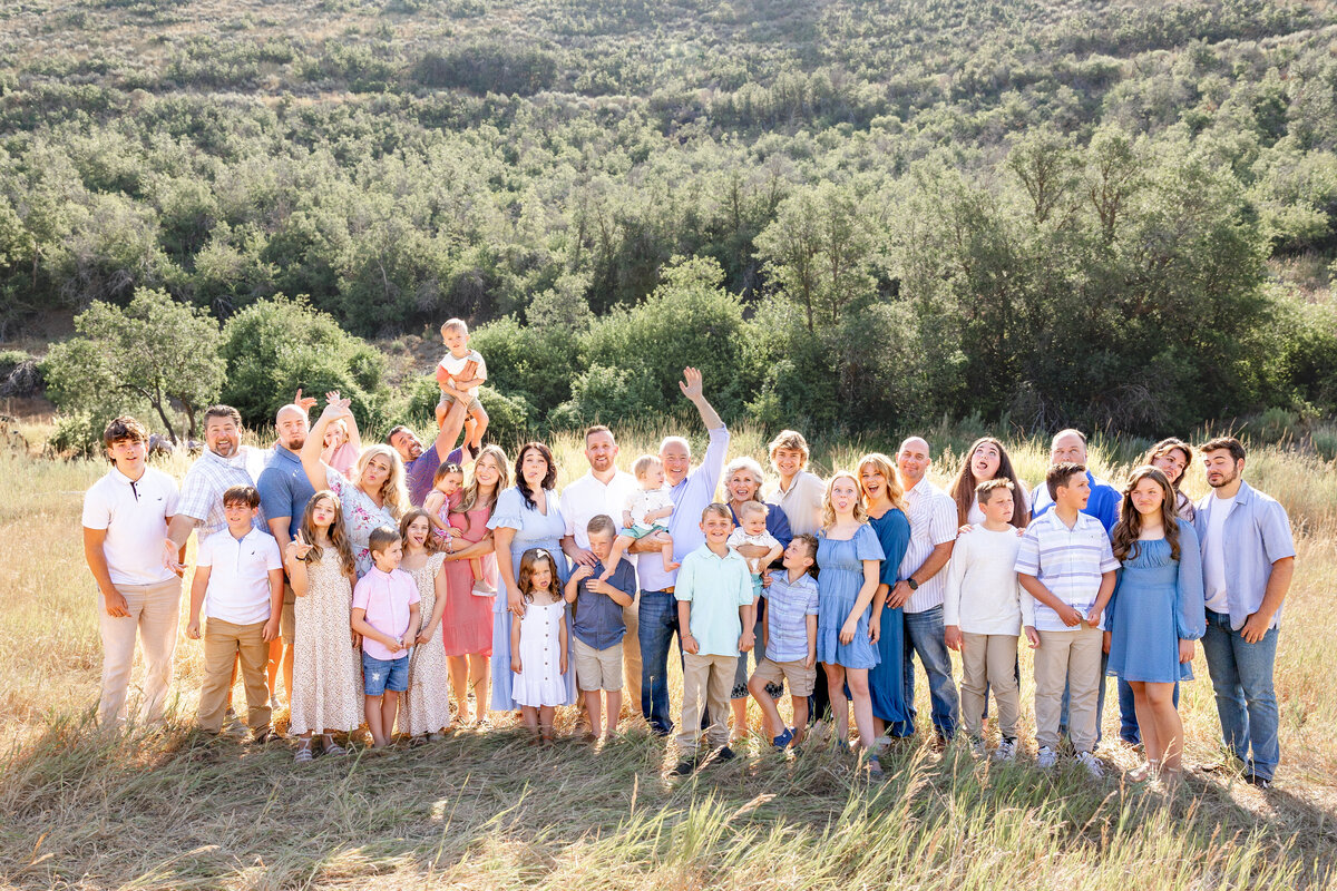A big family enjoys a lively group photo in a sunlit field, with laughter and movement bringing energy to the scene. A child is lifted into the air, adding a playful touch to this warm family reunion.