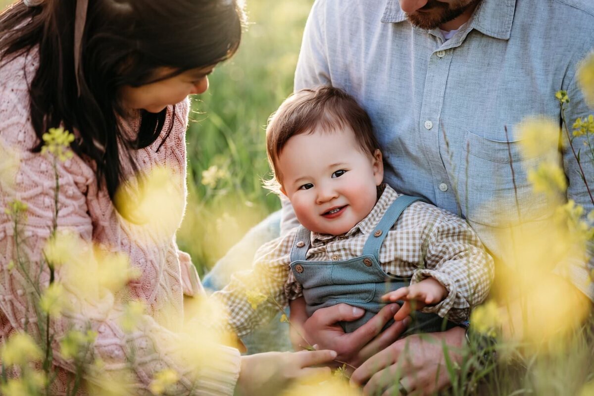baby boy smiles at camera. there are wildflowers behind him.