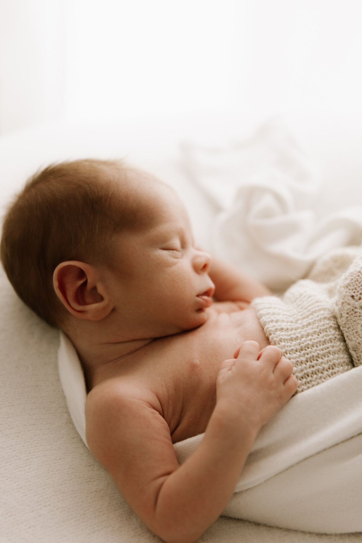 A side profile of a sleeping newborn swaddled in soft, cream-toned fabrics. The baby's gentle features, tiny hand, and textured knit pants are softly lit, evoking warmth, stillness, and a sense of safety.