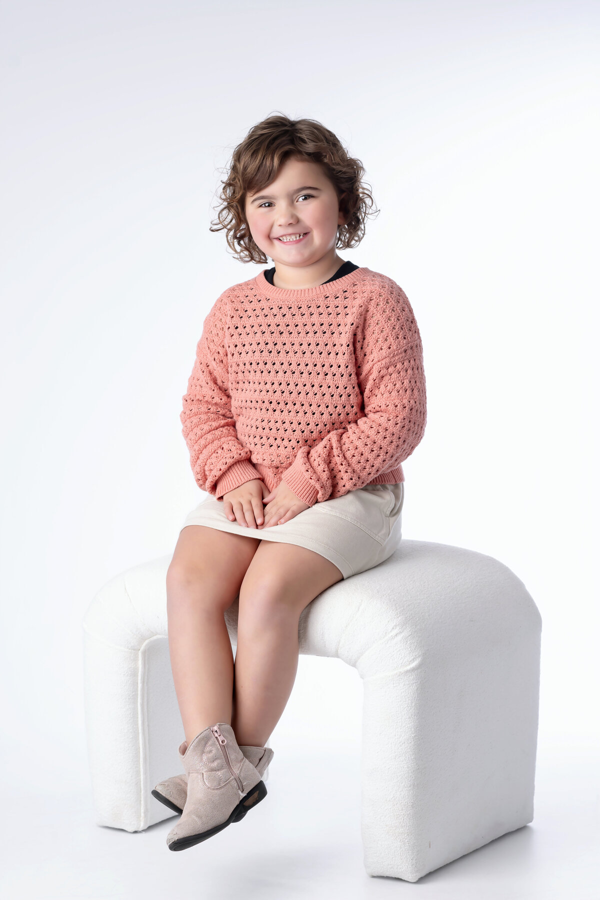 Young girl smiles as she sits on a white seat in a white studio