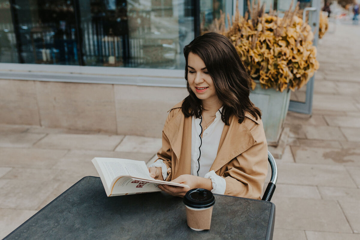 Sarah Berneche sits at an outdoor coffee table reading Body Respect. She is wearing a beige trench coat, a white silk scalloped shirt, and a gold coin necklace. Her shoulder-length brown hair rests at her shoulders. 