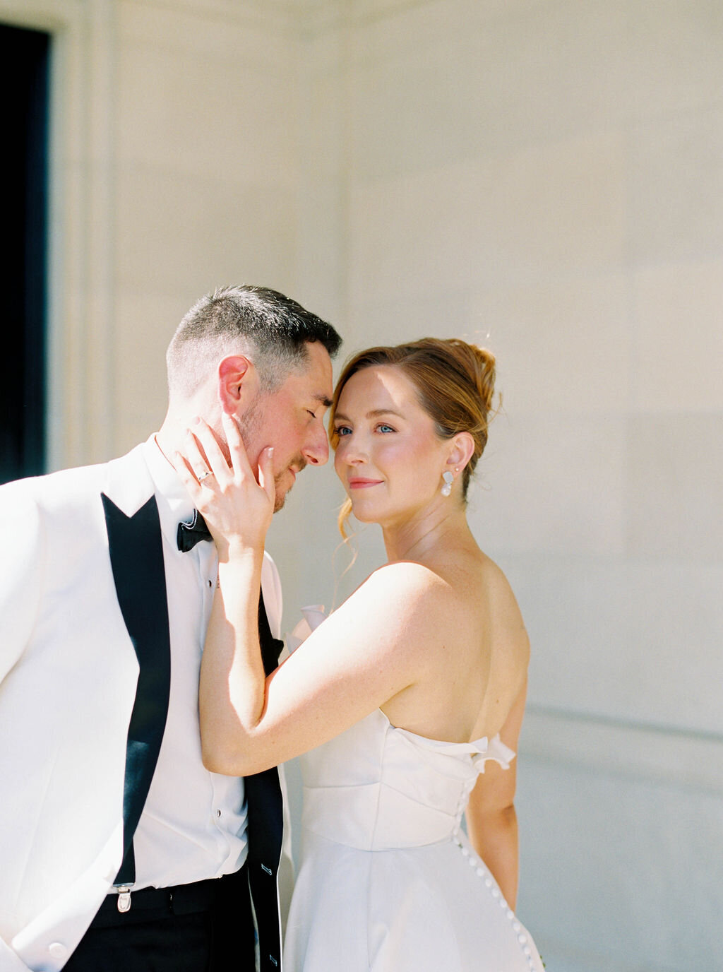 Rhode Island Elopement | A bride and groom share an intimate moment. She wears a strapless white dress, touching his face tenderly. He wears a white tuxedo with a black lapel.