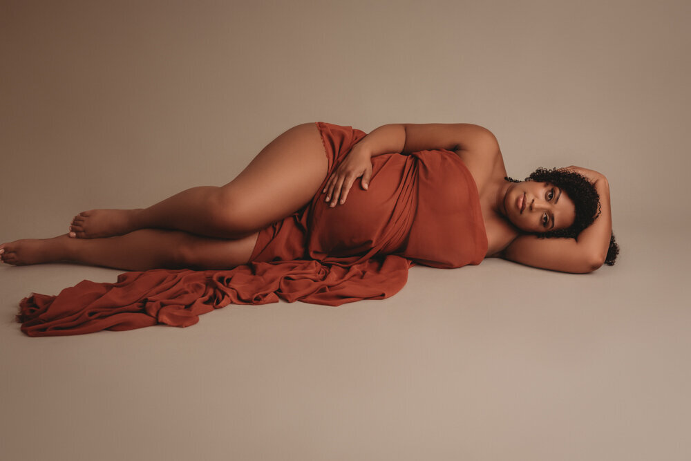 Atlanta, Georgia maternity photographer session in studio with pregnant woman laying on her side draped in burnt orange chiffon fabric holding her tummy.