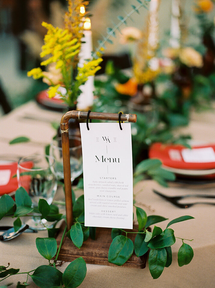 White dinner menu with black font hangs on a brass and wooden place card holder atop a table set with green garland.