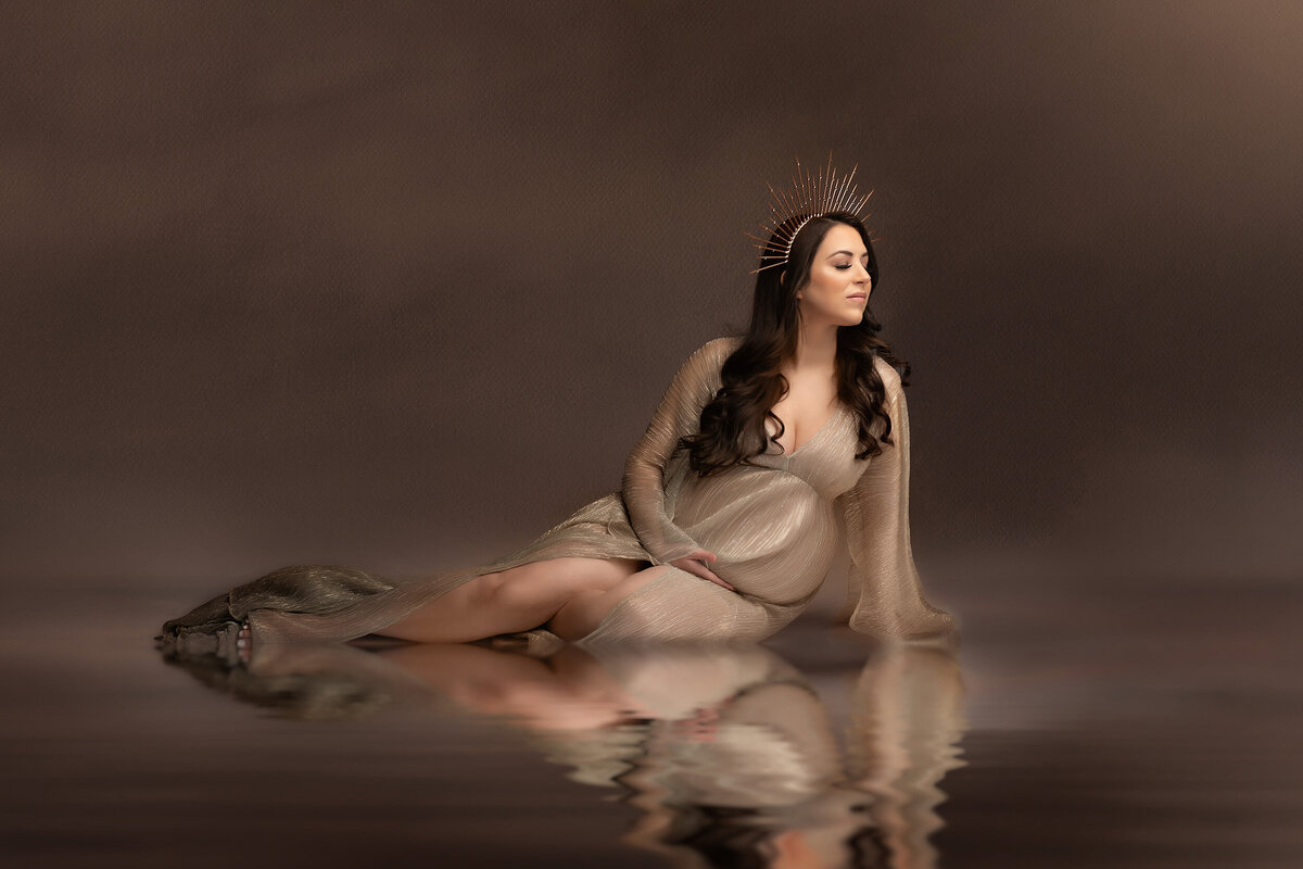 Pregnant woman sits mermaid style in dark pool of water reflecting her image. She wears a neutral, sheer maternity gown and a gold, spiked crown.