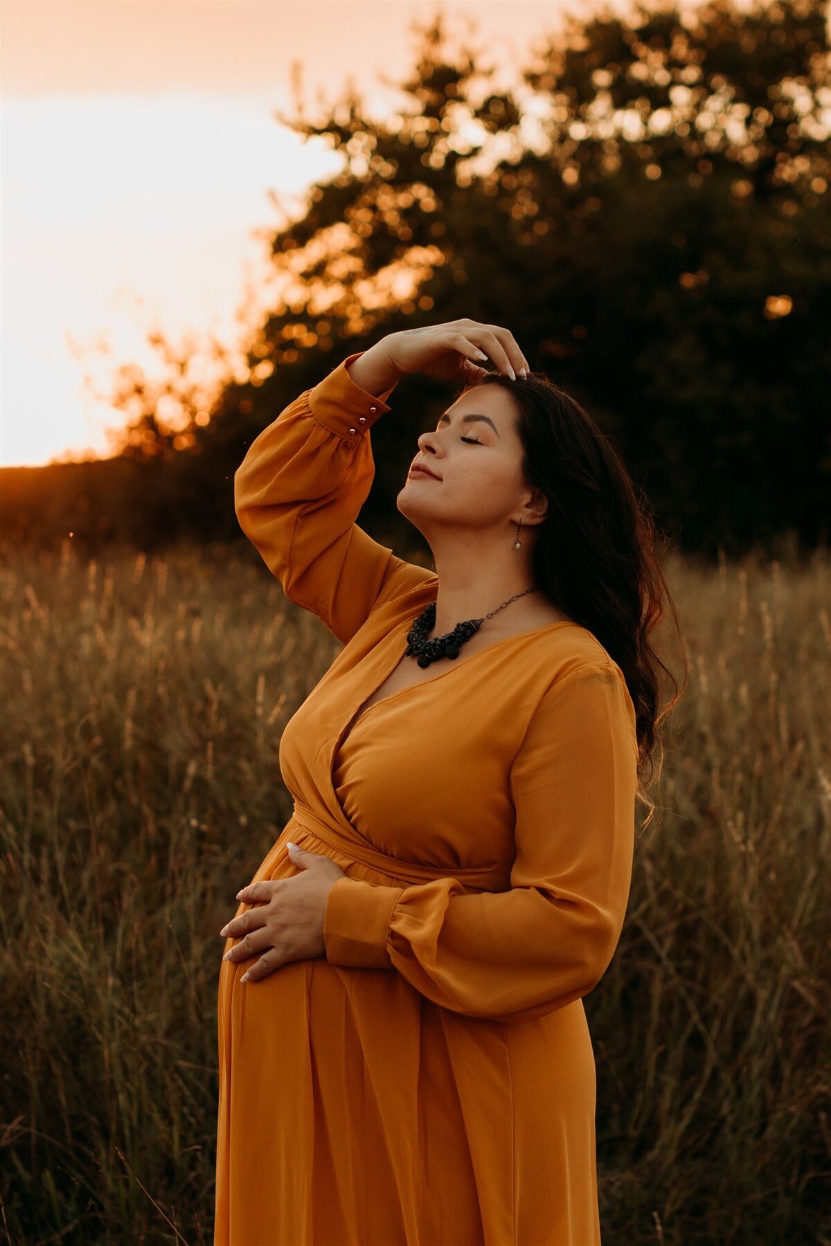 Pregnant woman in orange dress stands in front of tall grass and moves hand through her hair