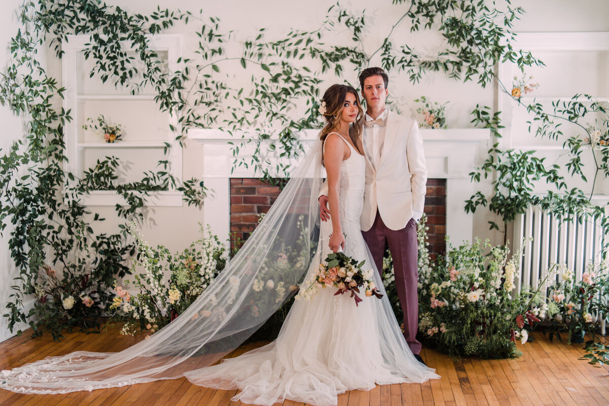 Bride wearing Alexandra Grecco standing with groom in white & rose tuxedo surrounded by vines and flowers after wedding ceremony
