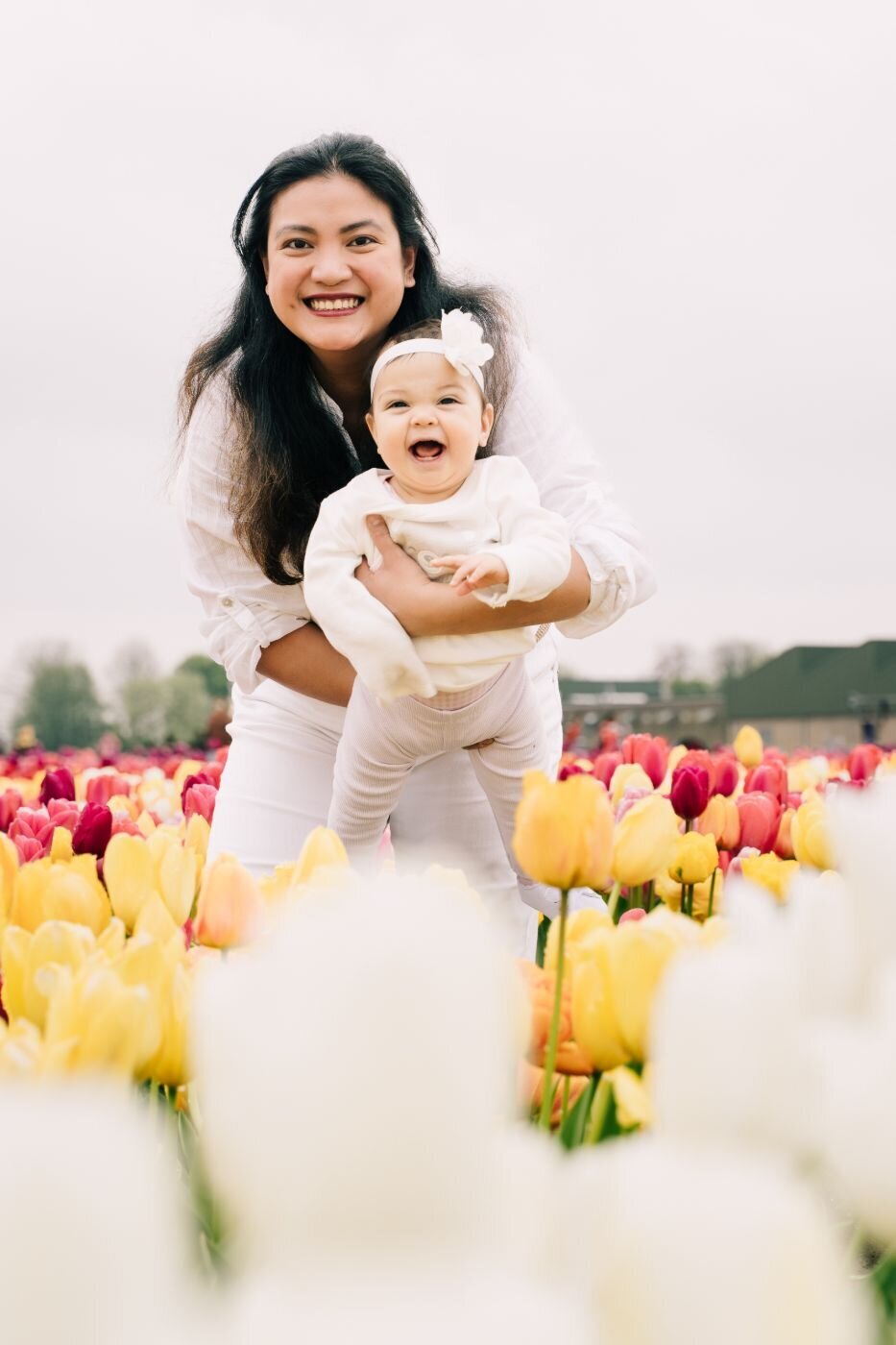 Mother in white holding her smiling baby among vibrant rows of tulips in the Netherlands.