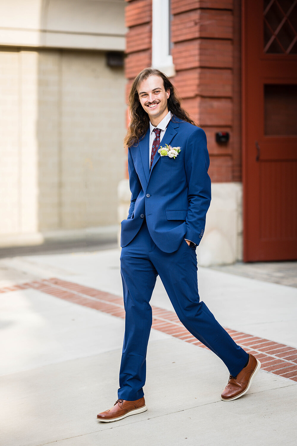 A groom in a navy suit walks with his hands in his pockets and smiles towards something behind the photographer (not photographed) on his elopement day in Downtown Roanoke.