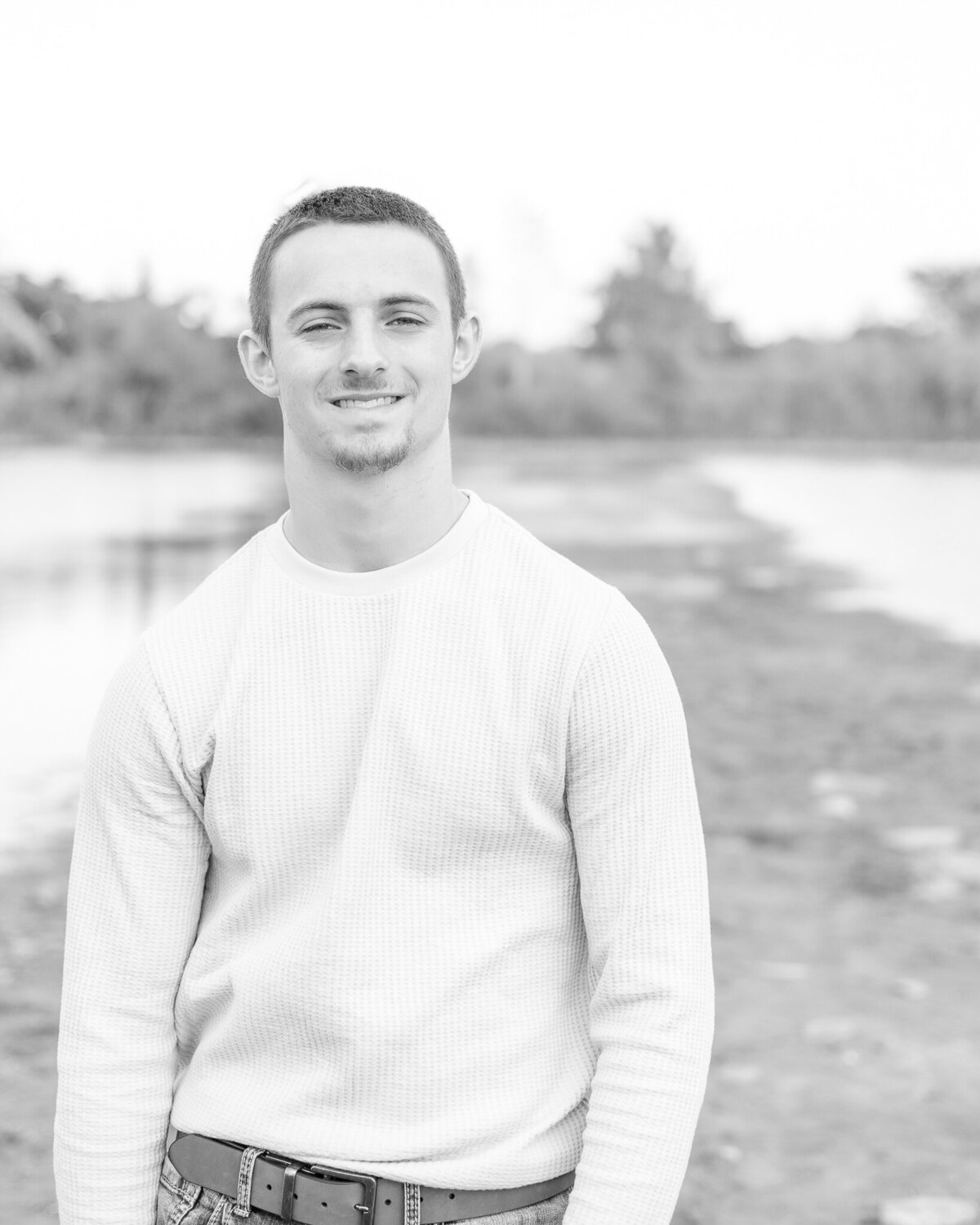 black and white photo of a high school senior male with water in the background, looking at the camera smiling, wearing a long sleeve shirt