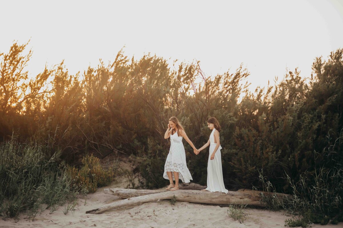 Mother holds teenage daughters hand while walking across a log on the beach, both wearing white flowy dresses