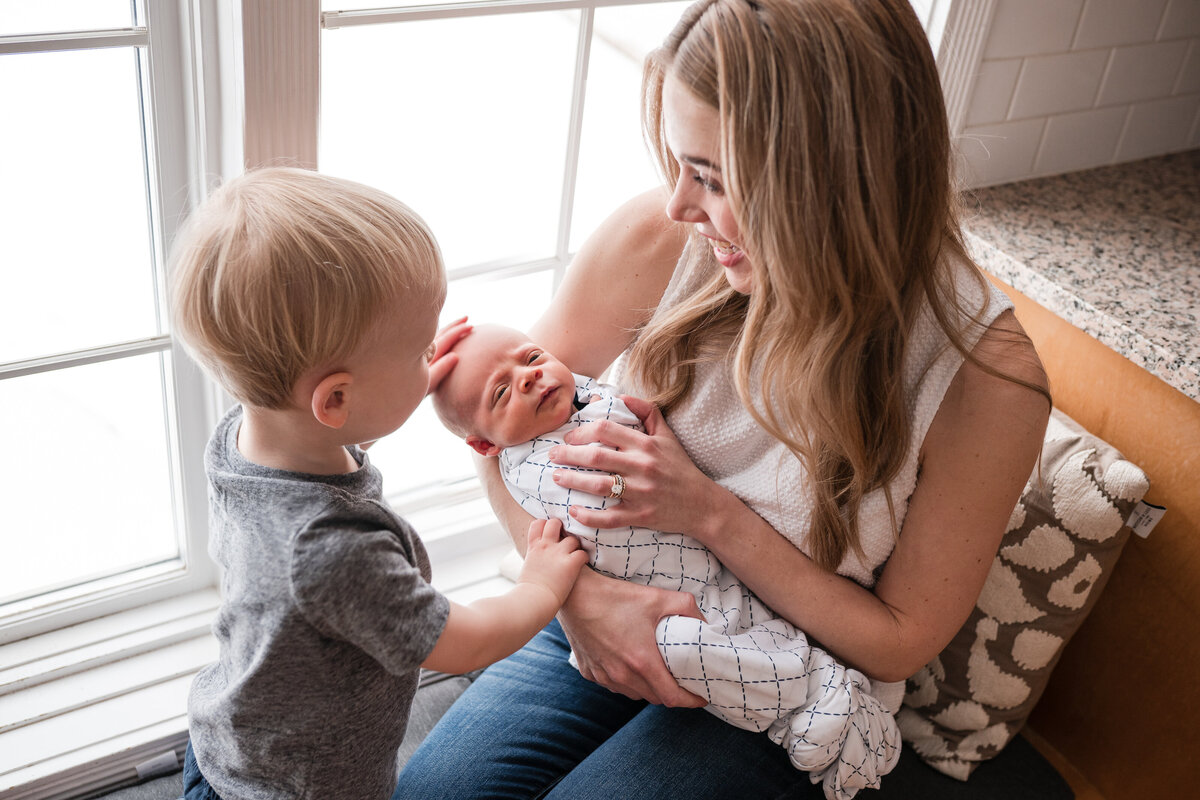 A toddler gently pats the head of a newborn baby who is swaddled and resting in their mother's arms by a window, both mother and child are smiling.