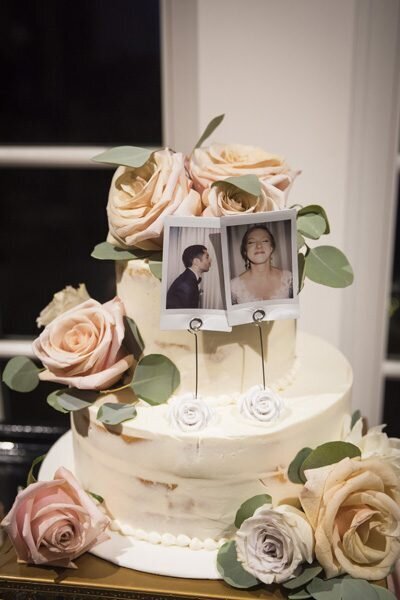 A two-tier wedding cake with white icing, pastel-colored roses and a Polaroid of the groom and one of the bride making silly faces.