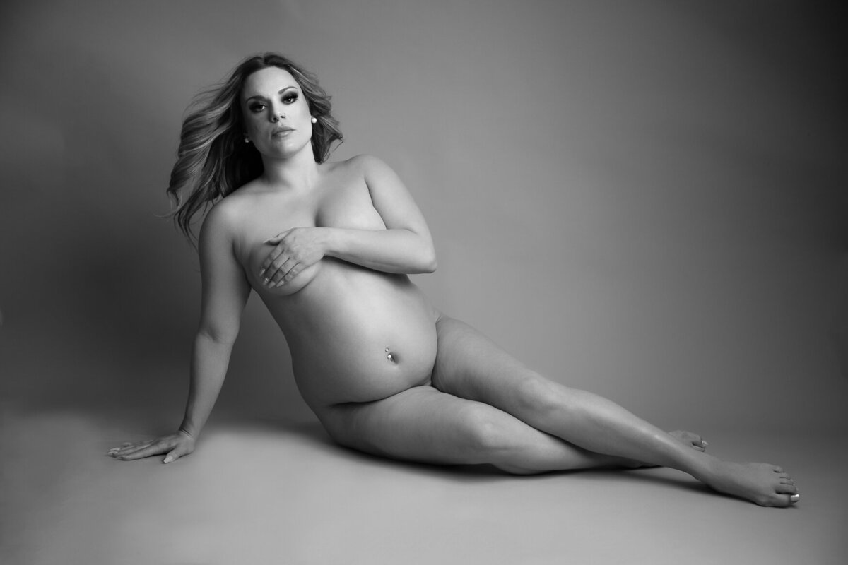 A pregnant woman laying down on the floor posing to the camera nude and covering her boobs with her hand