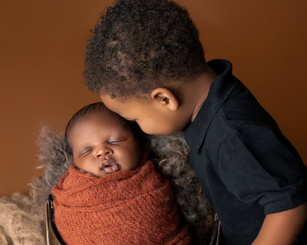 Older brother leaning in to kiss newborn baby swaddled in rust wrap with dark orange background | Newborn sibling portrait in Cedar Park and Austin