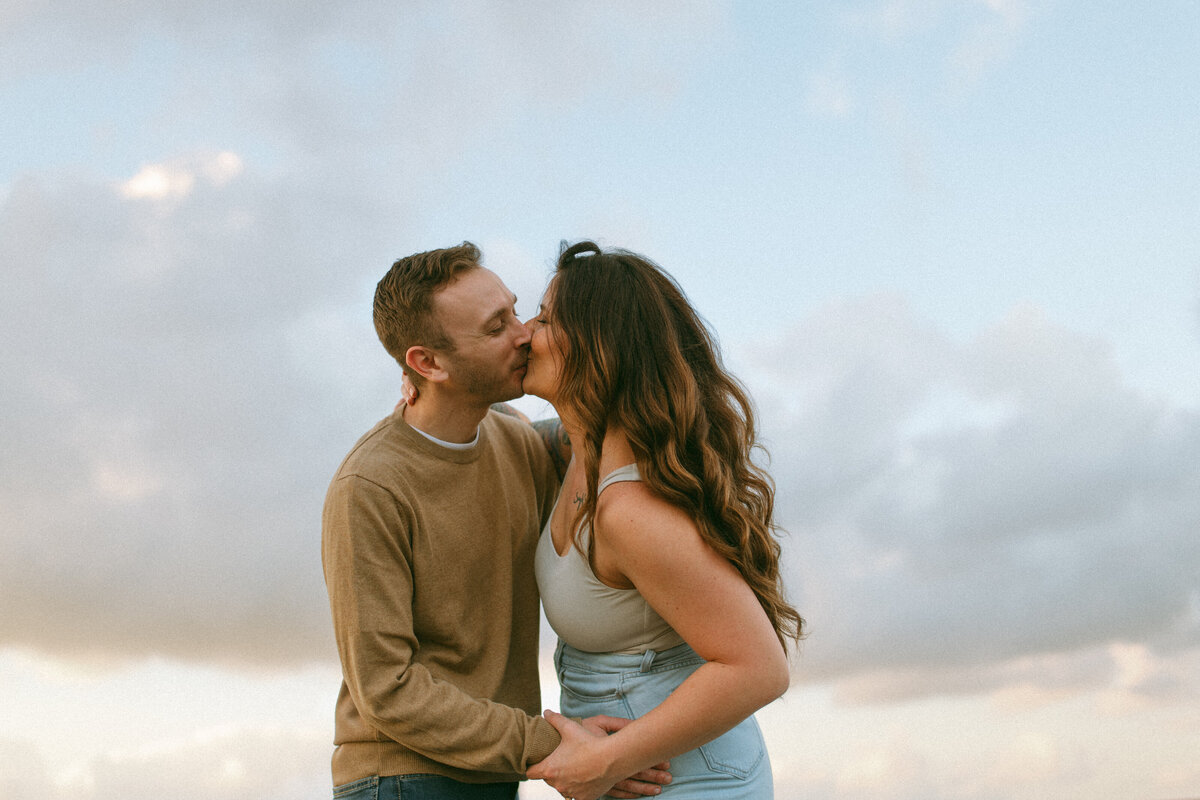 a couple kissing with the cotton candy colored sky above them.
