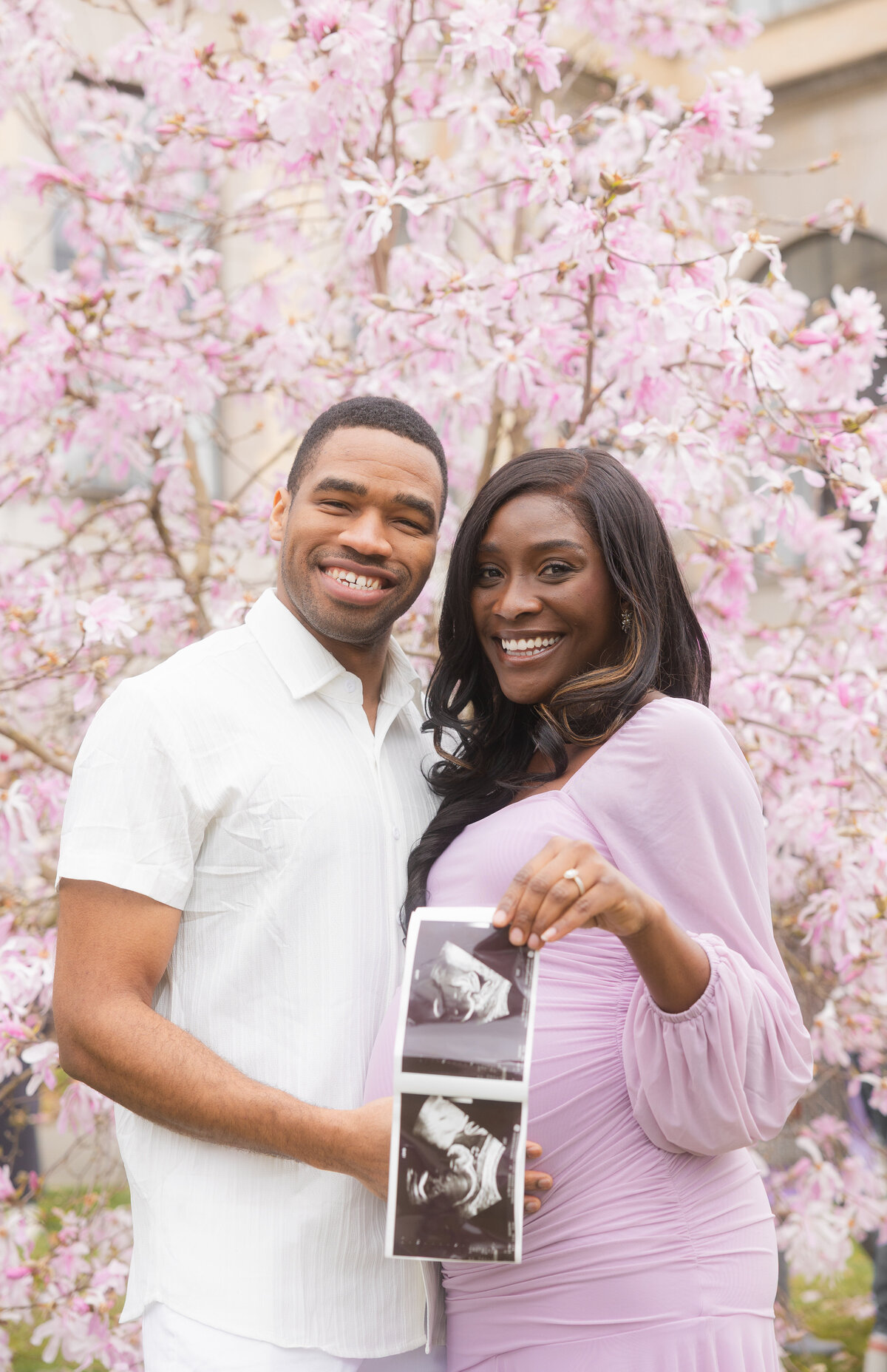 A joyful moment as the couple smiles at each other, holding up their ultrasound scan in front of the vibrant cherry blossoms at the University of Washington campus, celebrating their exciting news.