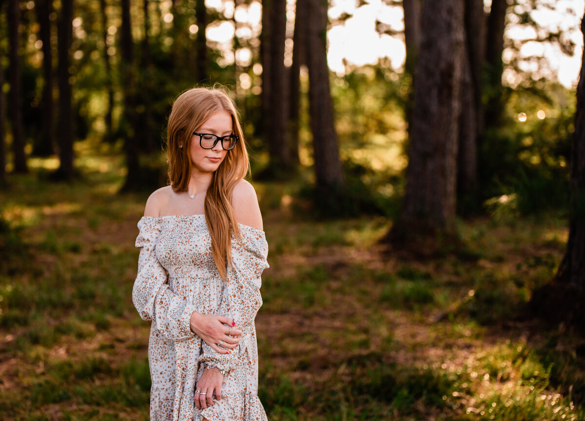 A senior in an off the shoulder dress looks over her shoulder in a forest like setting.