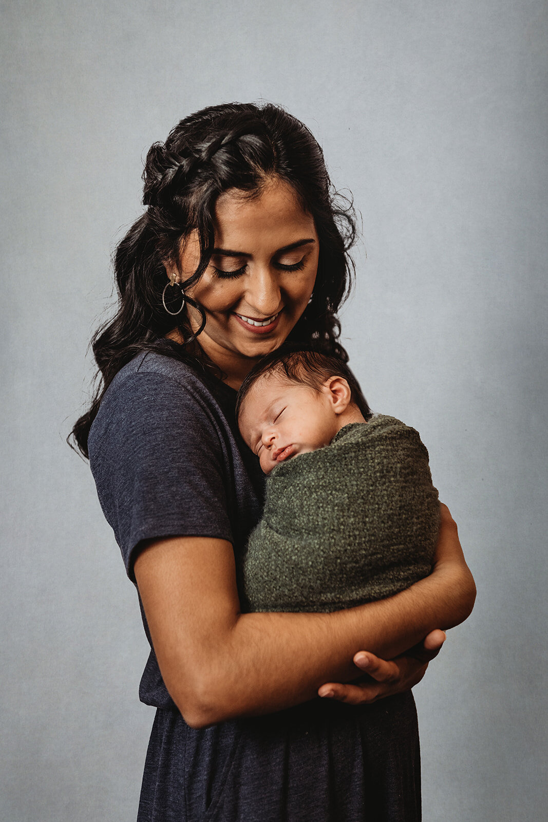 newborn photos with mother in grey dress holding her baby who is wrapped in a dark green swaddle for an in studio newborn session photographed by Family photographers Maryland