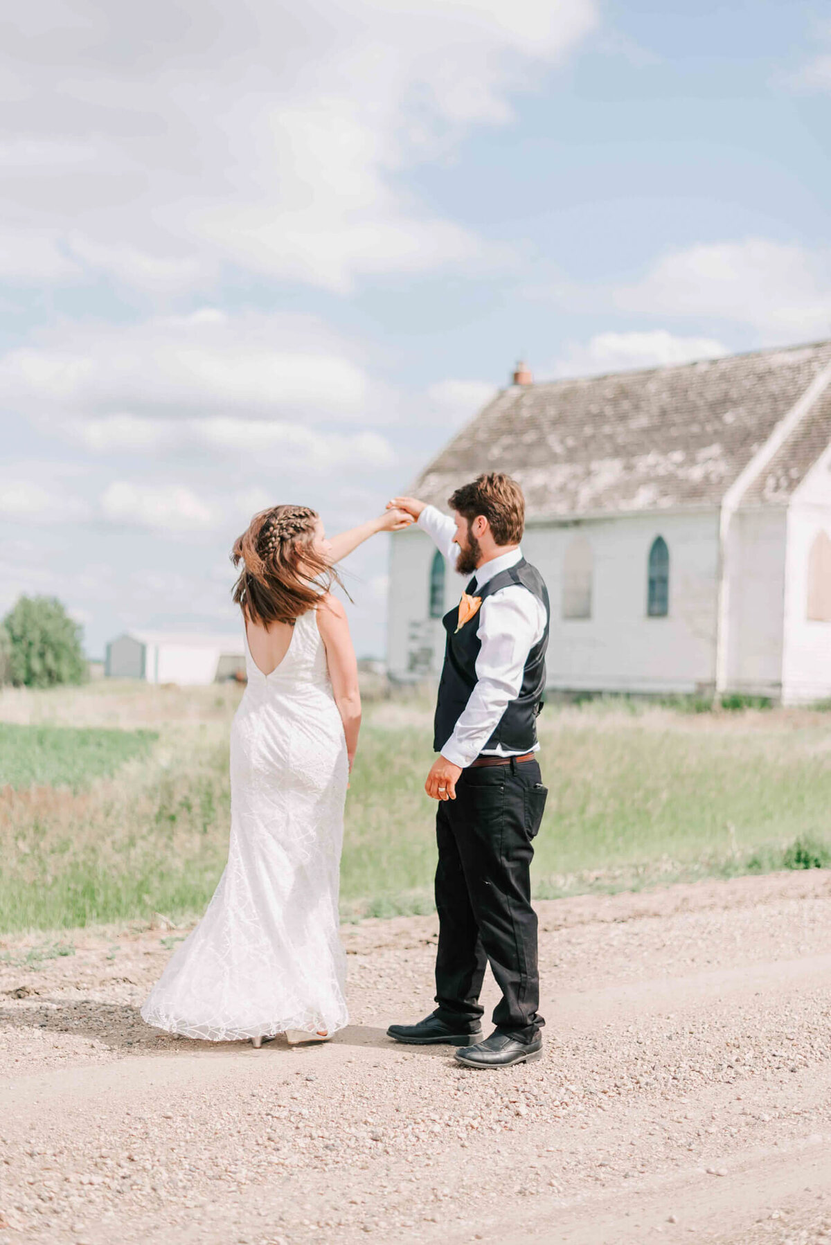 Groom is spinning bride while they dance on a gravel road.