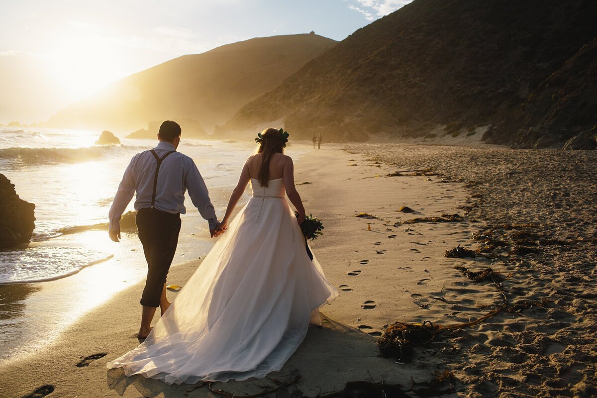 Bride and groom hold hands and walk down the beach during sunset