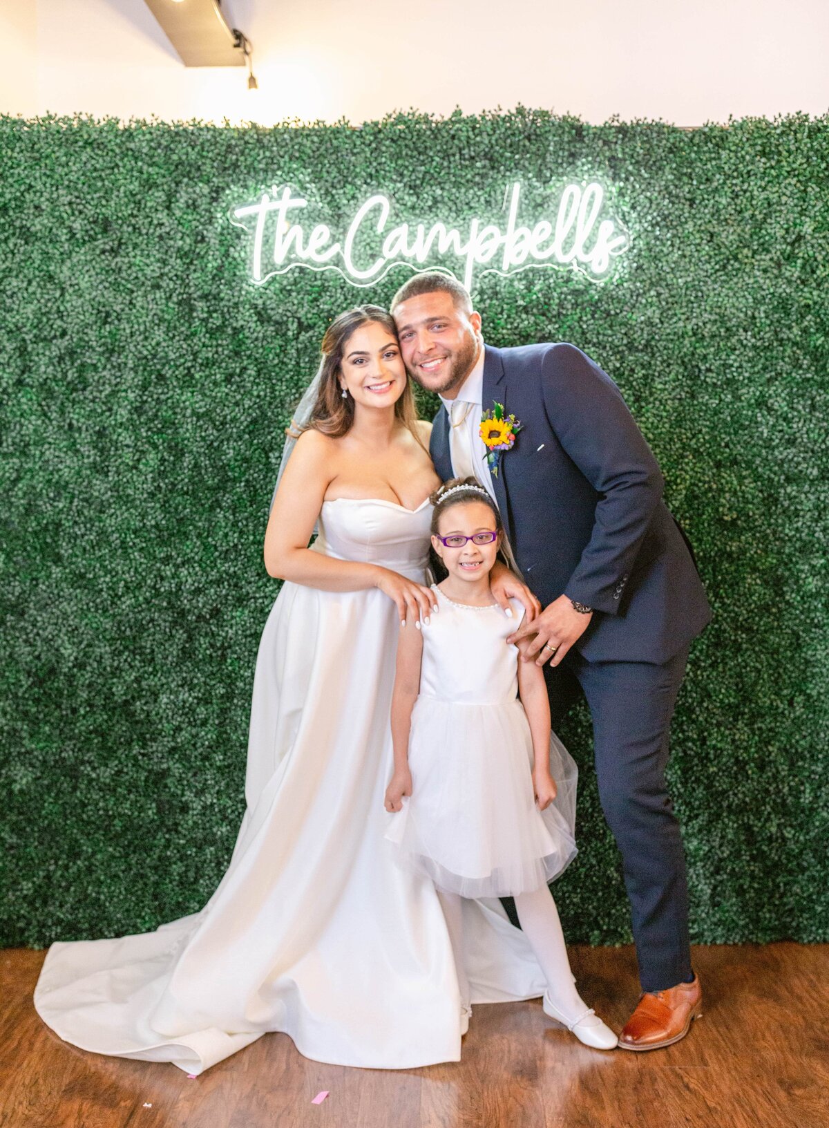 Bride, groom and daughter standing under Neon last name sign during wedding reception at the Refinery at 120 in Culpeper, Virginia. Captured by Bethany Aubre Photography.