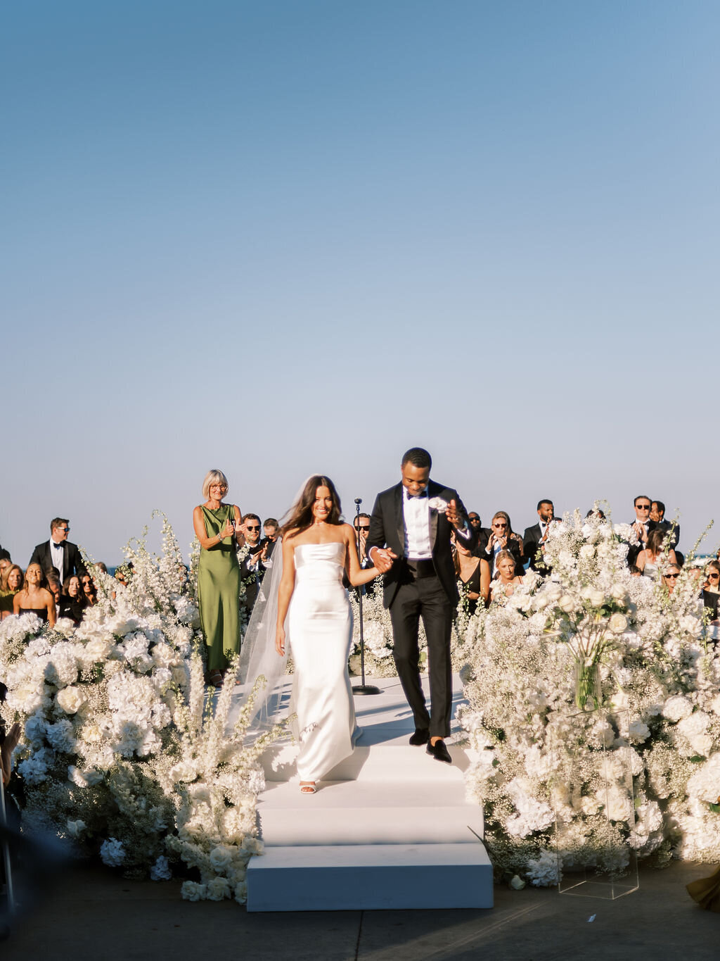 The newlyweds walk down the aisle as guests cheer, marking the start of their new journey together.
