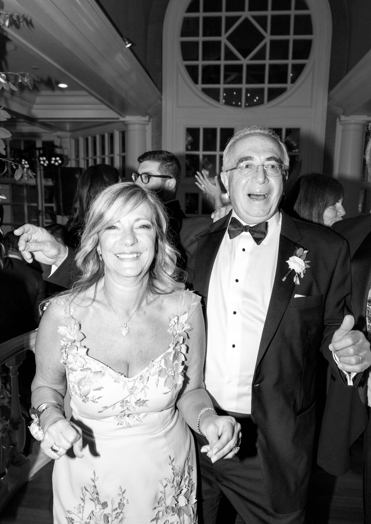 A black and white photo of a joyful couple at an event. The woman is wearing a floral dress, while the man is dressed in a tuxedo. Both are smiling and appear to be enjoying themselves in a lively atmosphere.