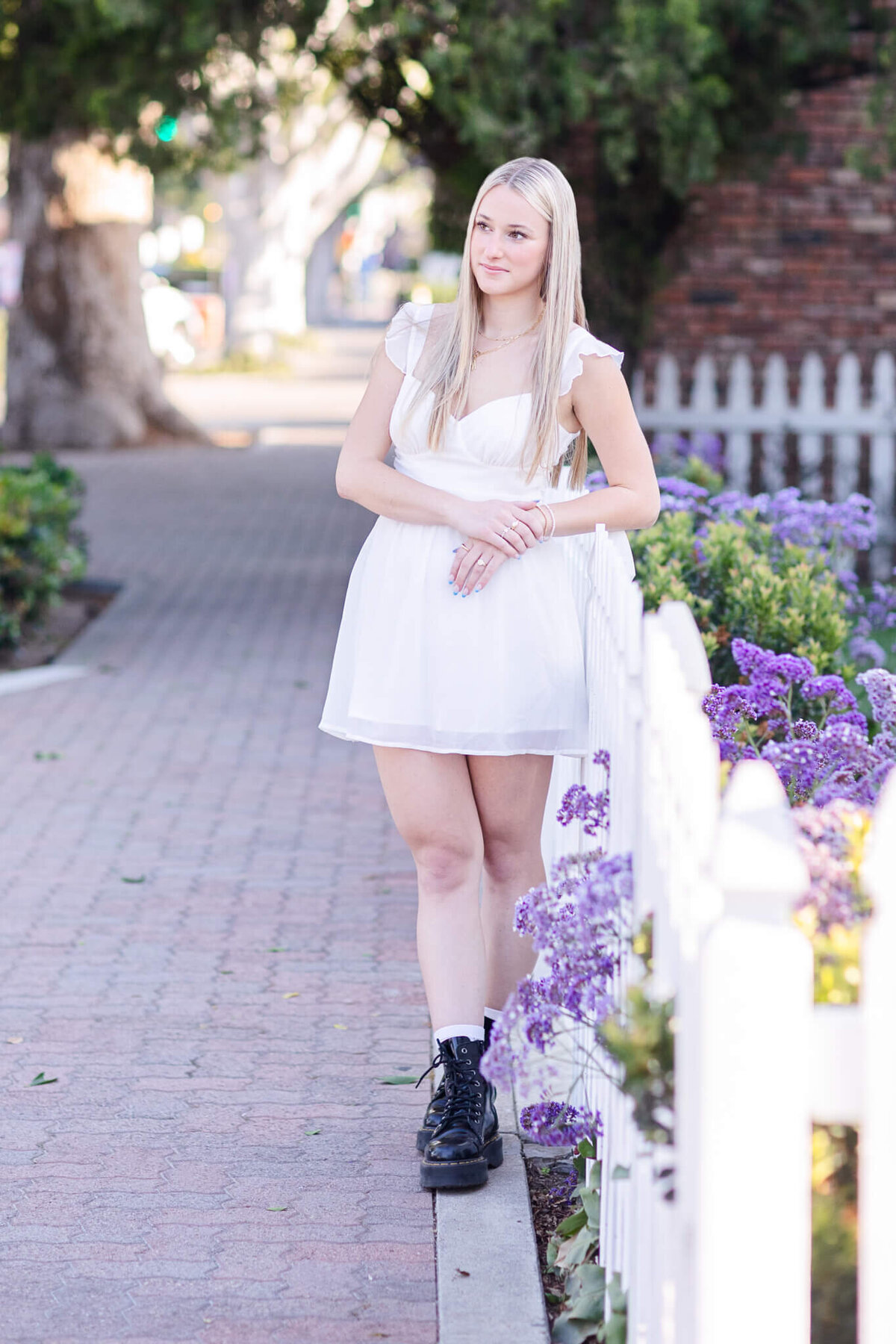 A young woman with long blonde hair wearing a white dress and black boots stands on a sidewalk lined with purple flowers and a white picket fence. She is outdoors, with trees and a brick building in the background.