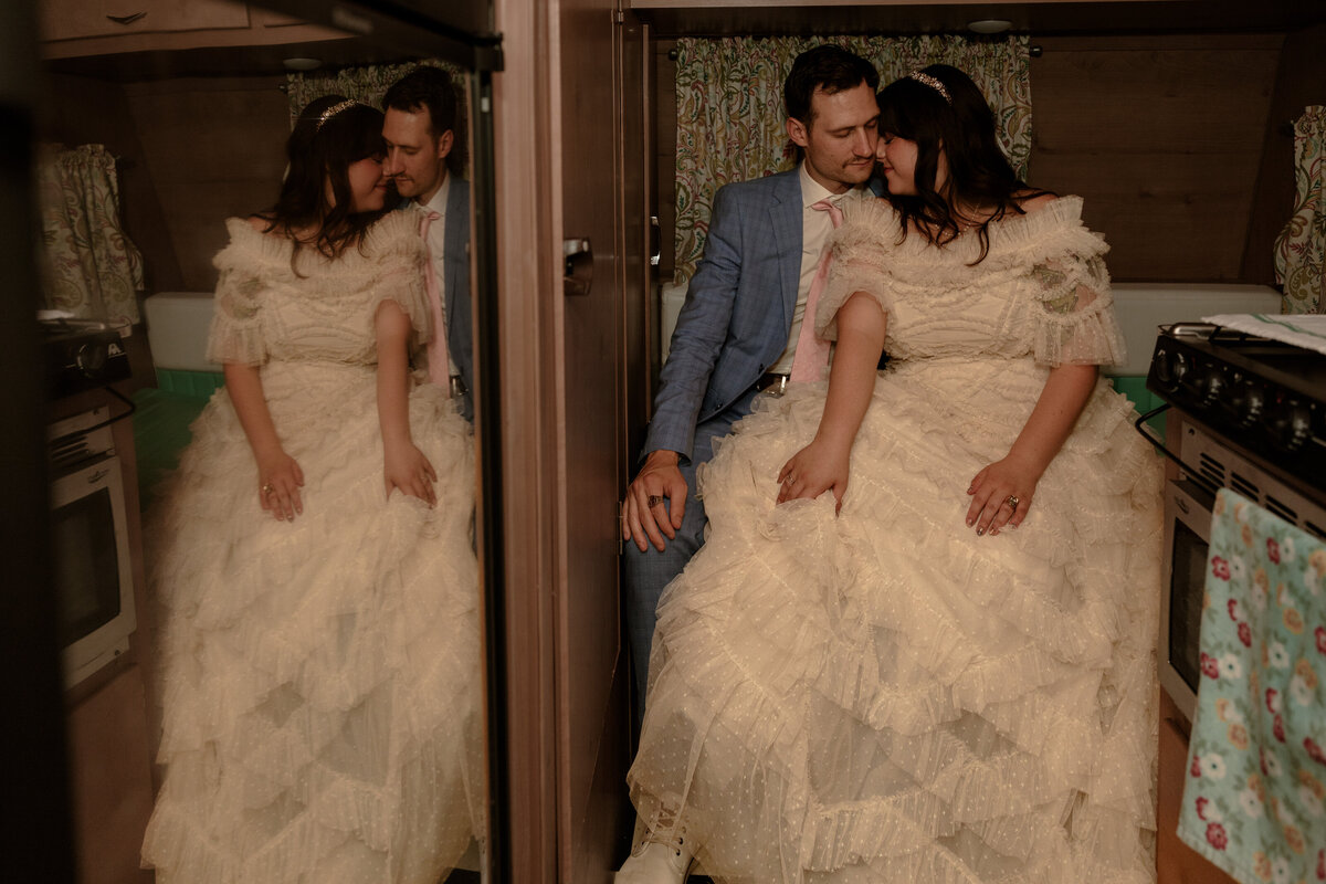 Bride and groom sit closely together in a vintage camper, their foreheads touching with eyes closed, reflected in the mirrored door beside them.