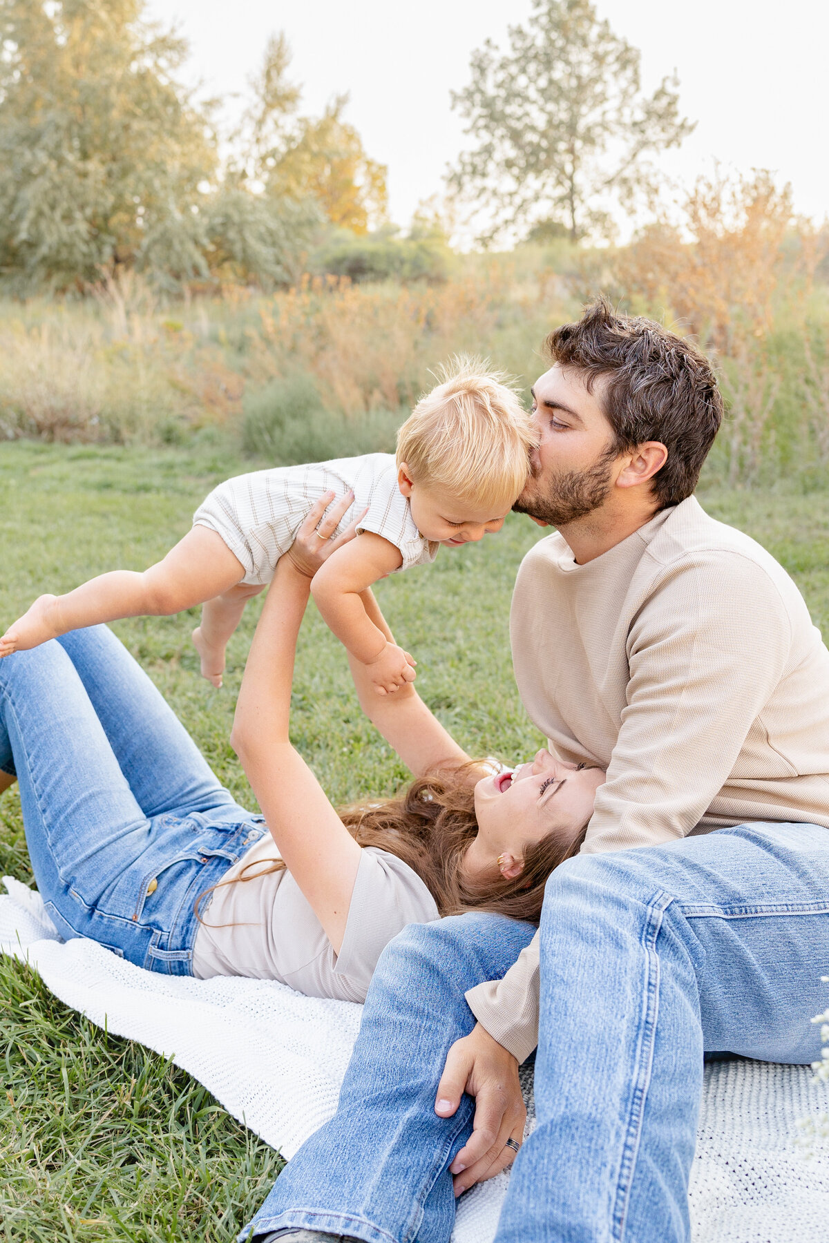 A father kisses his baby on the head as the mother lifts him into the air, capturing a sweet and candid family moment in the scenic outdoors of Eagle Mountain, Utah. Taken by Cassie Lee Photography