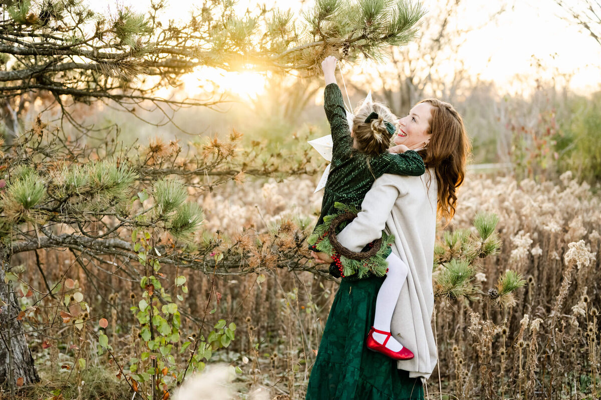 Mom lifting daughter to place ornament on pine tree at a sunset family session  near Chicago,  IL.