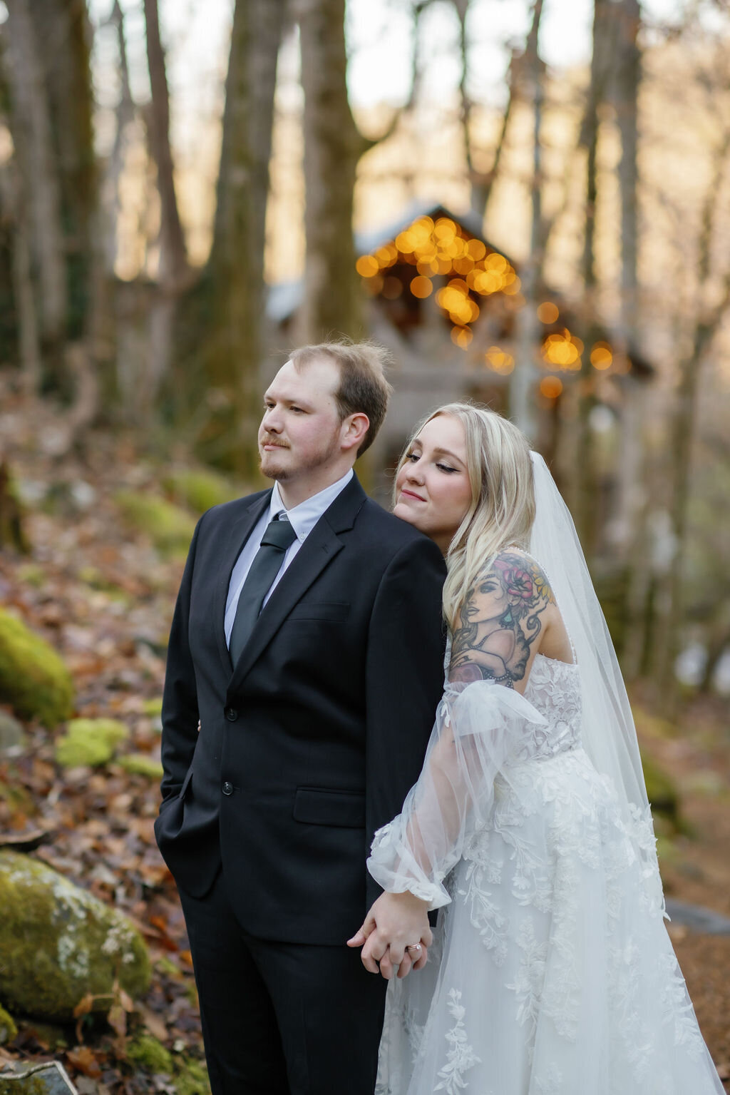 Ely's Mill elopement picture with bride standing behind her groom and holding his chest with the pavilion in the distance through the woods lit with twinkle lights for Gatlinburg elopement 