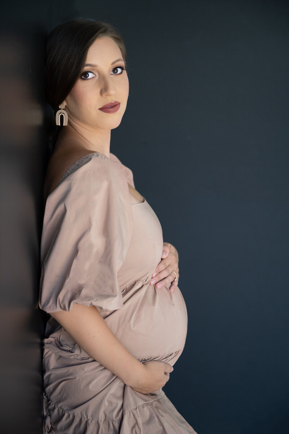 A young woman posing against a black wall wearing a soft nude dress showing her pregnant belly