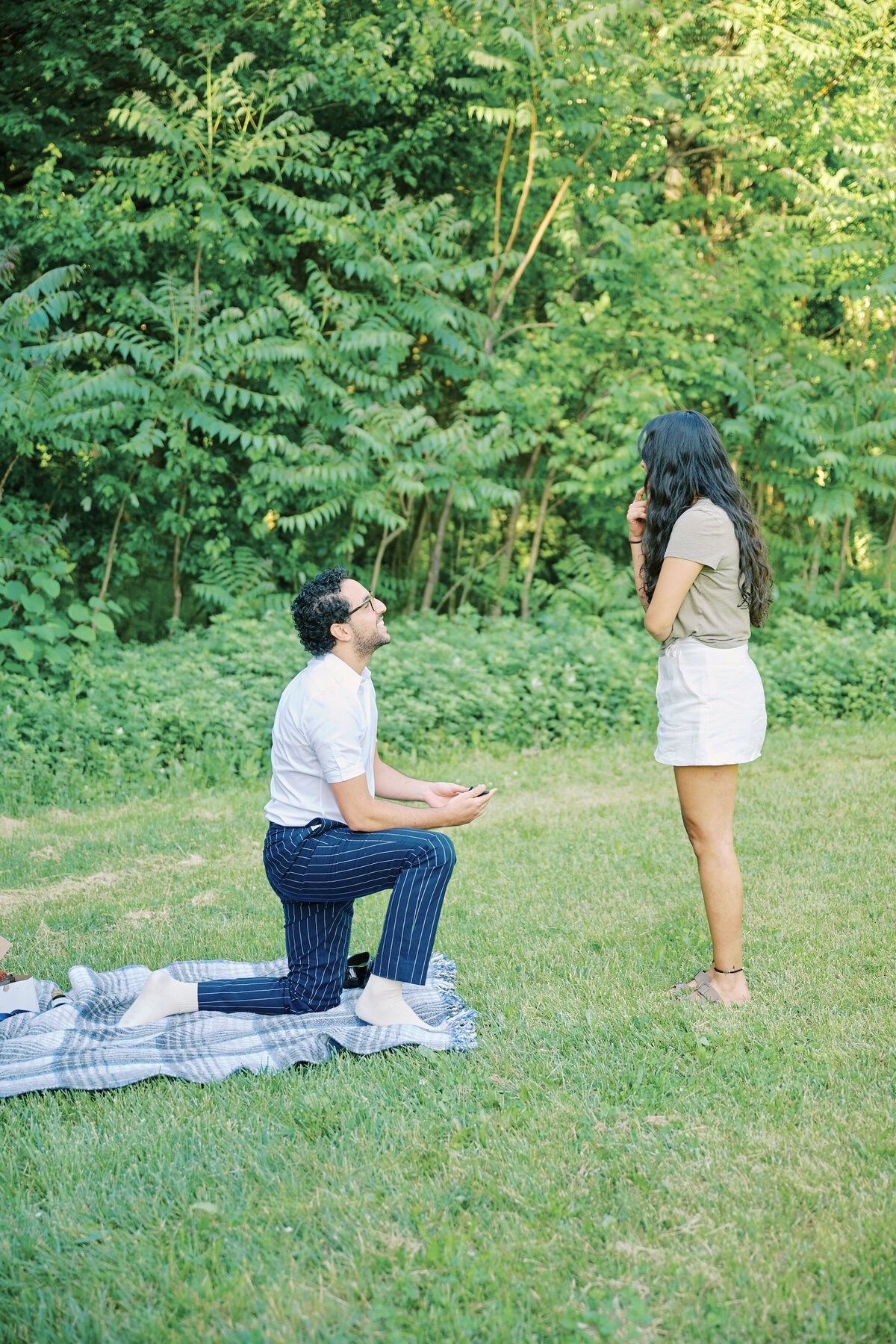 A man is down on bended knee proposing.  Picnic Proposal in New York City, Queens.  New York City Proposal Photography, Corey Lamar Photography