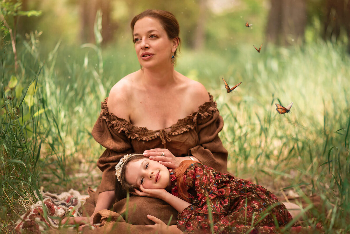 Brittany danielle captures a stunning portrait of a mother and daughter. Mother is sitting on the ground in the grass while her young daughter rests her head in her lap while smiling at the camera. Mother em braces her while looking off into the distance. They are surrounded by butterflies.