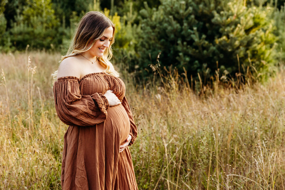 woman expecting her first baby  looking at her baby bump and thinking about how her life is about to change as she stands in a field at sunset captured by Ashley Kalbus Photography