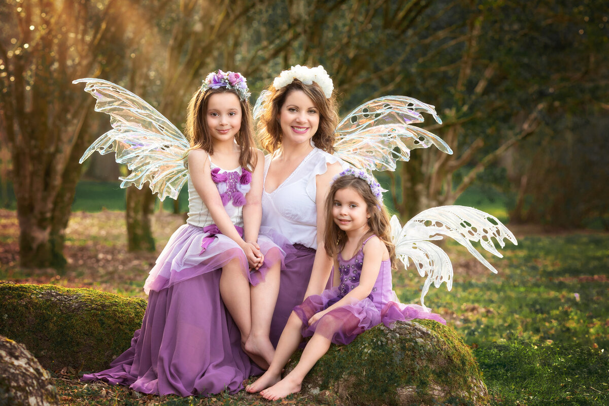 Mom and 2 daughters dressed as fairies in the park with trees and wildflowers.  All 3 are wearing fairy wings from Crone Designs.  They are all dressed in white and purple.  They are sitting on boulders and rocks at Jungle Gardens in Avery Island.