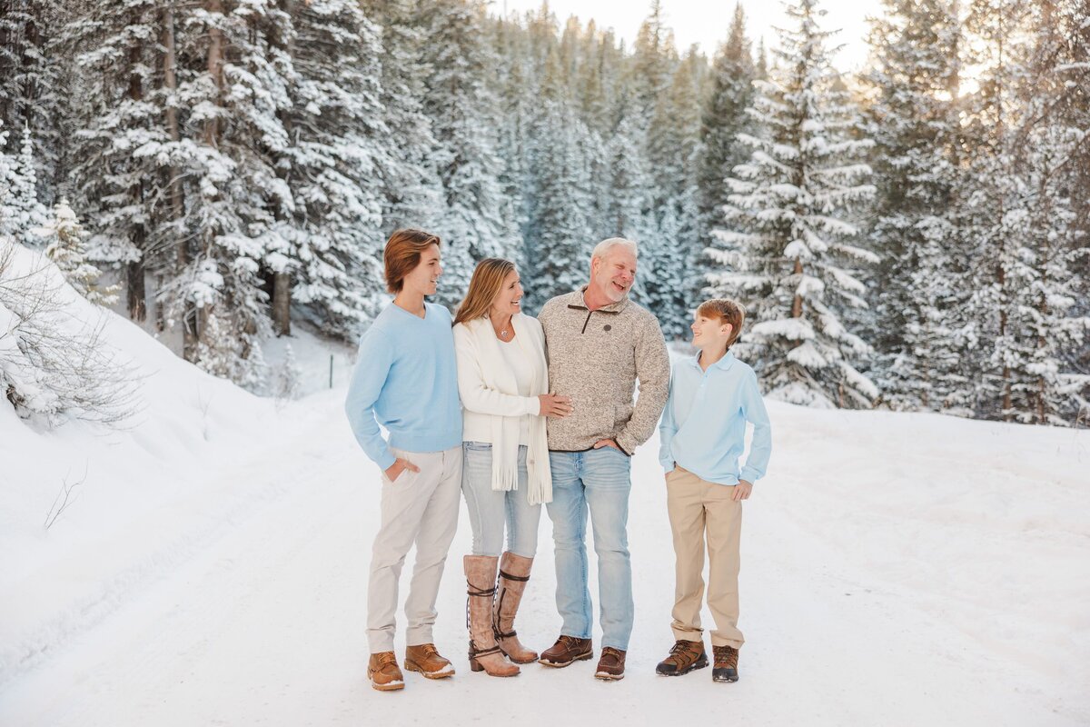 A mom dad and teen laugh while looking towards the younger son in a snowy mountain