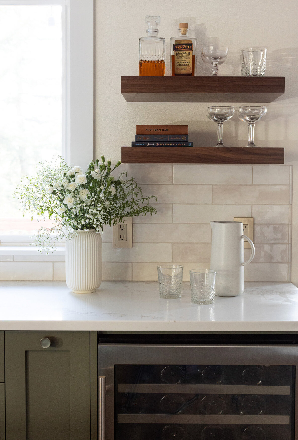 Closeup of a kitchen bar with a wine fridge, green kitchen cabinets, white countertop, beige subway tile backsplash. A white vase of flowers sits on the counter. A white pitcher and two glasses sit on the counter. On the wall are two walnut floating shelves with alcohol, glasses, and books.