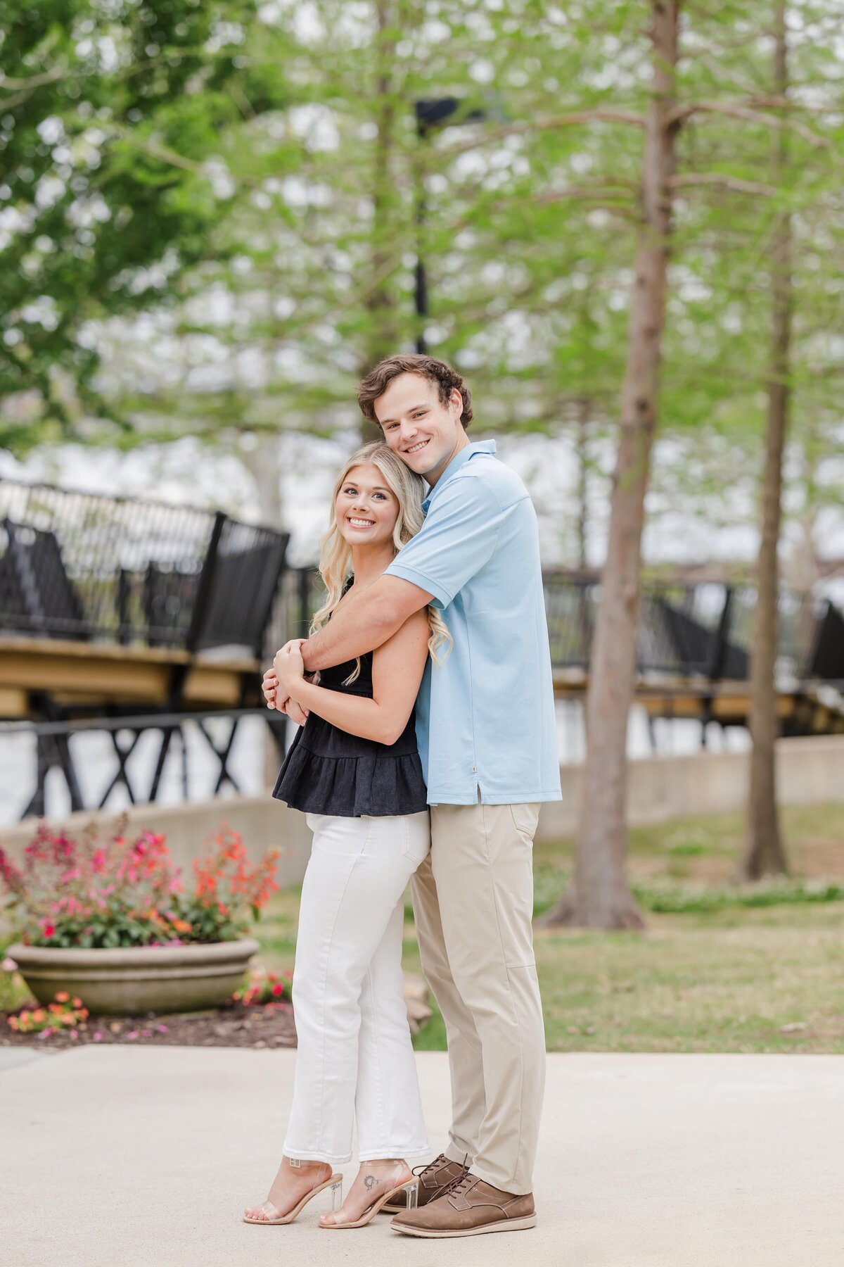 A man hugs his fiancee from behind while standing in a park sidewalk