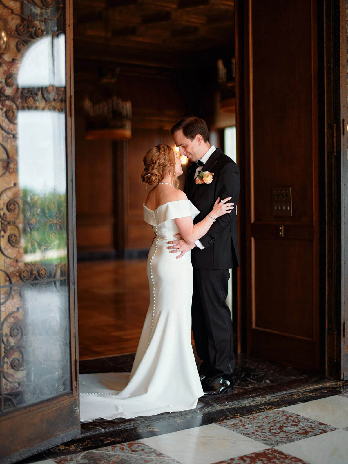 A bride in an off-the-shoulder white gown and a groom in a black suit embrace and smile at each other, beautifully captured by a film photographer NJ, inside an elegant, warmly lit venue with wooden details and patterned floors.