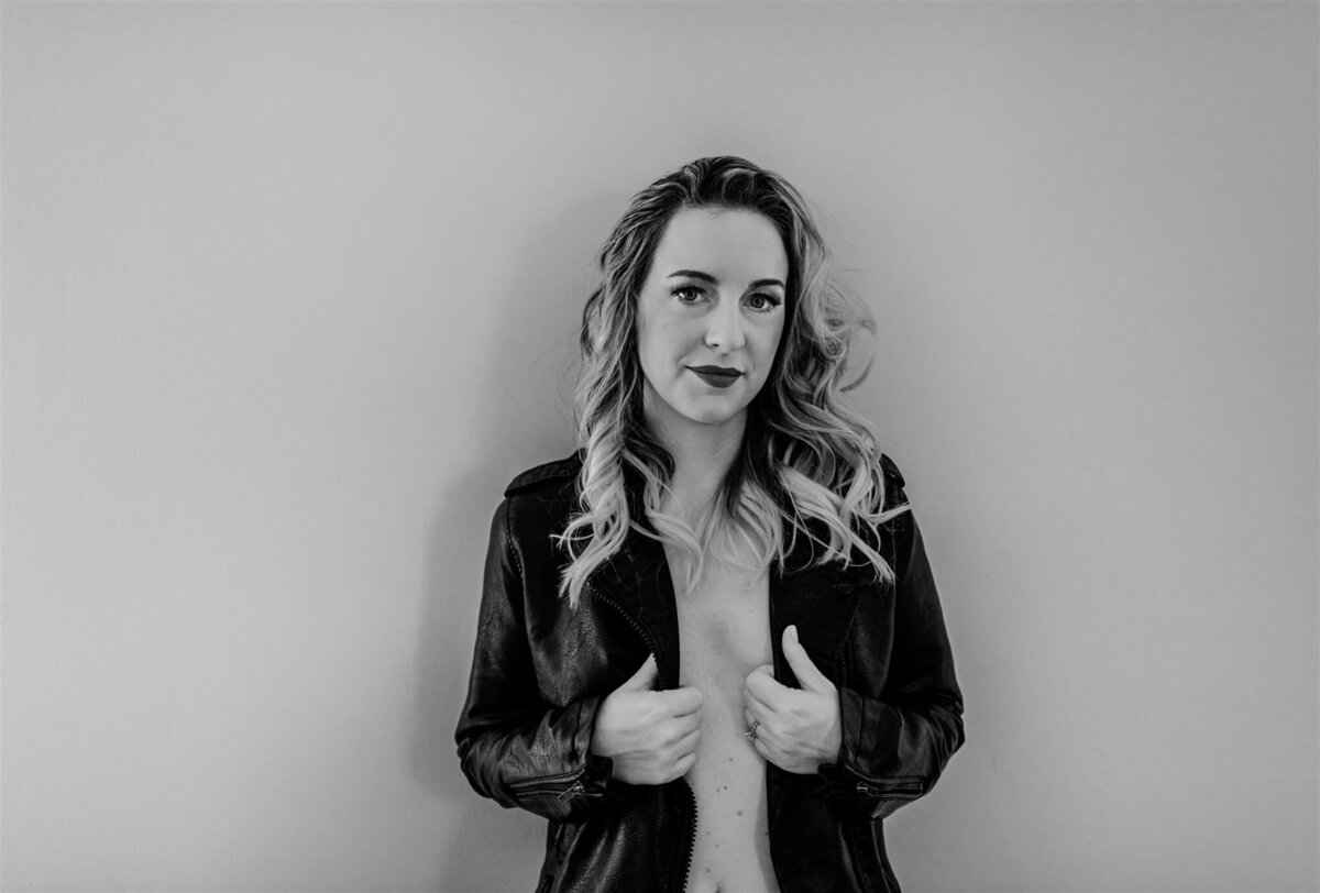 woman wearing nother but a black leather jacket leans against a white wall while holding her jacket to barely cover her chest captured by Baltimore photographers