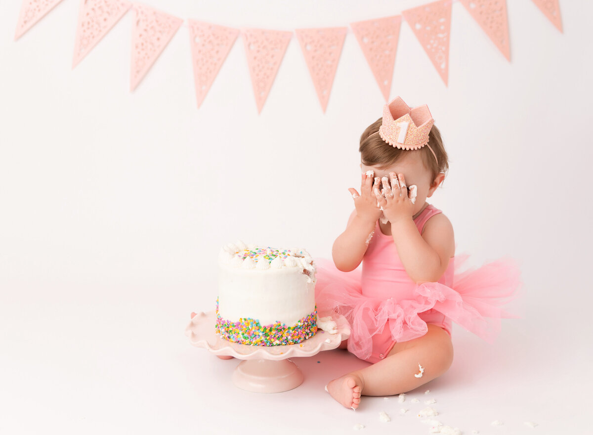 Baby girl in pink tutu with a white sprinkle cake sits for her first birthday cake smash photoshoot. Baby has icing on her hands and is playing peekaboo.