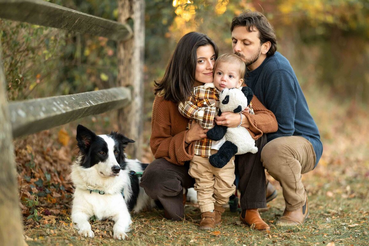 Fall family photo of mother, father, toddler, and dog next to rustic fence and autumn foliage