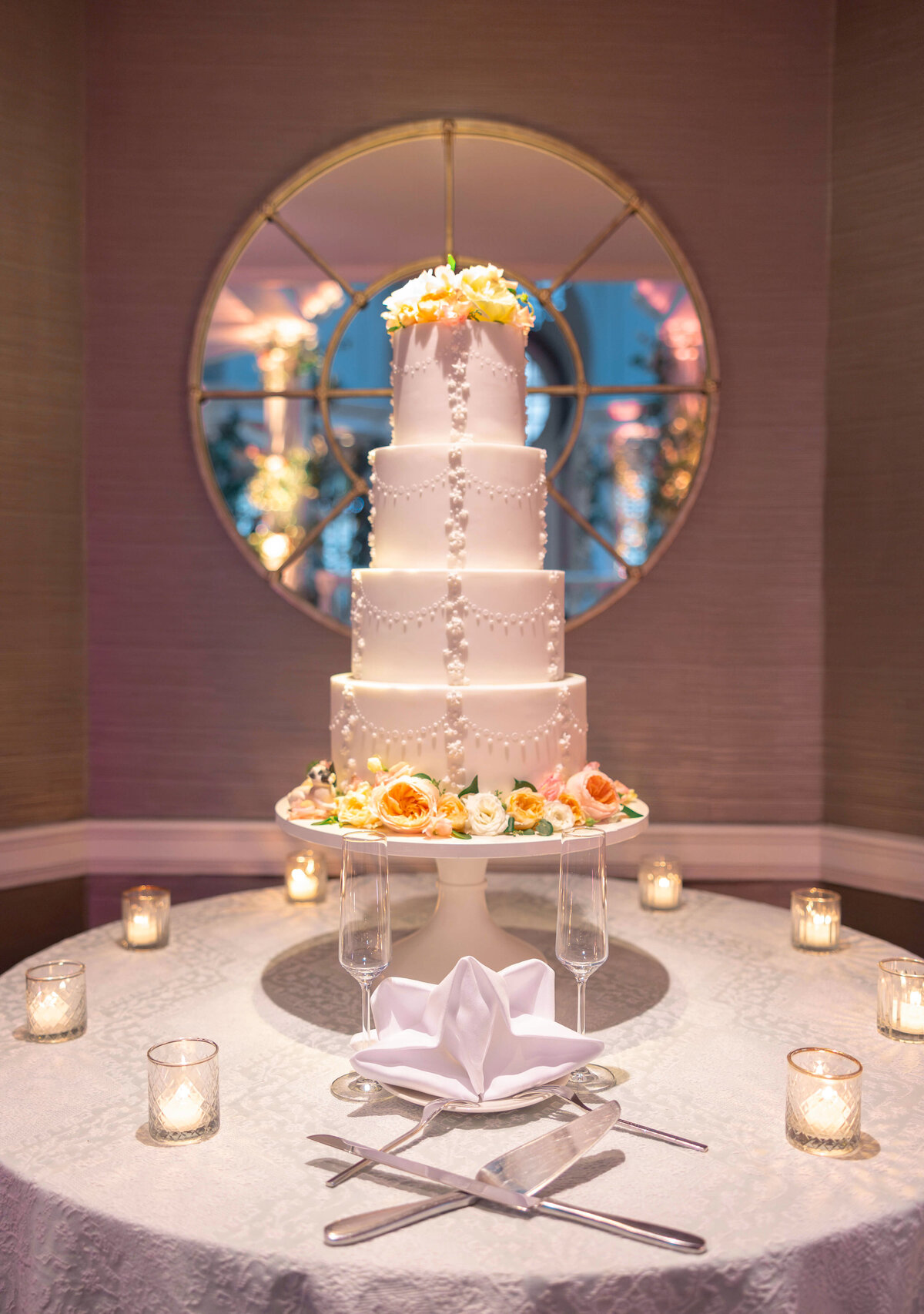 Elegant four-tiered white wedding cake adorned with pearls and topped with flowers, placed on a round table. Surrounded by decorative candles and flutes, with a folded napkin in the front. A large mirror reflects the rooms warm ambiance.