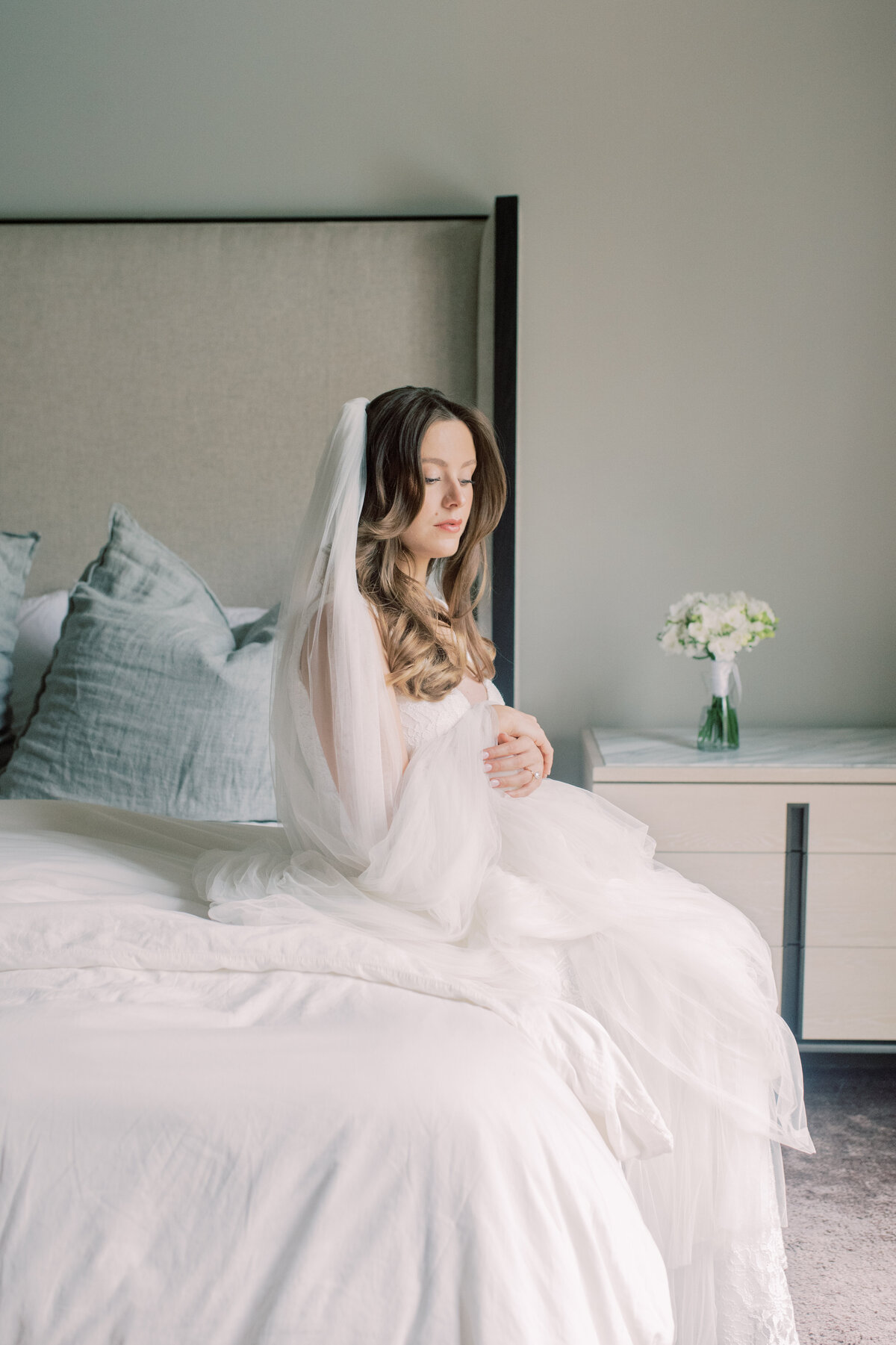 A serene photograph of a pregnant bride in a soft, natural light, gently holding her veil while seated on a bed, creating a calm and reflective atmosphere.
