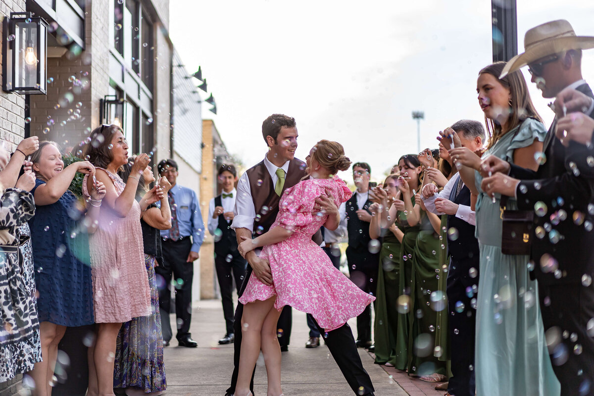A couple spins joyfully in the center of a cheering crowd outdoors, surrounded by people blowing bubbles and clapping, dressed in semi-formal attire on a bright day.