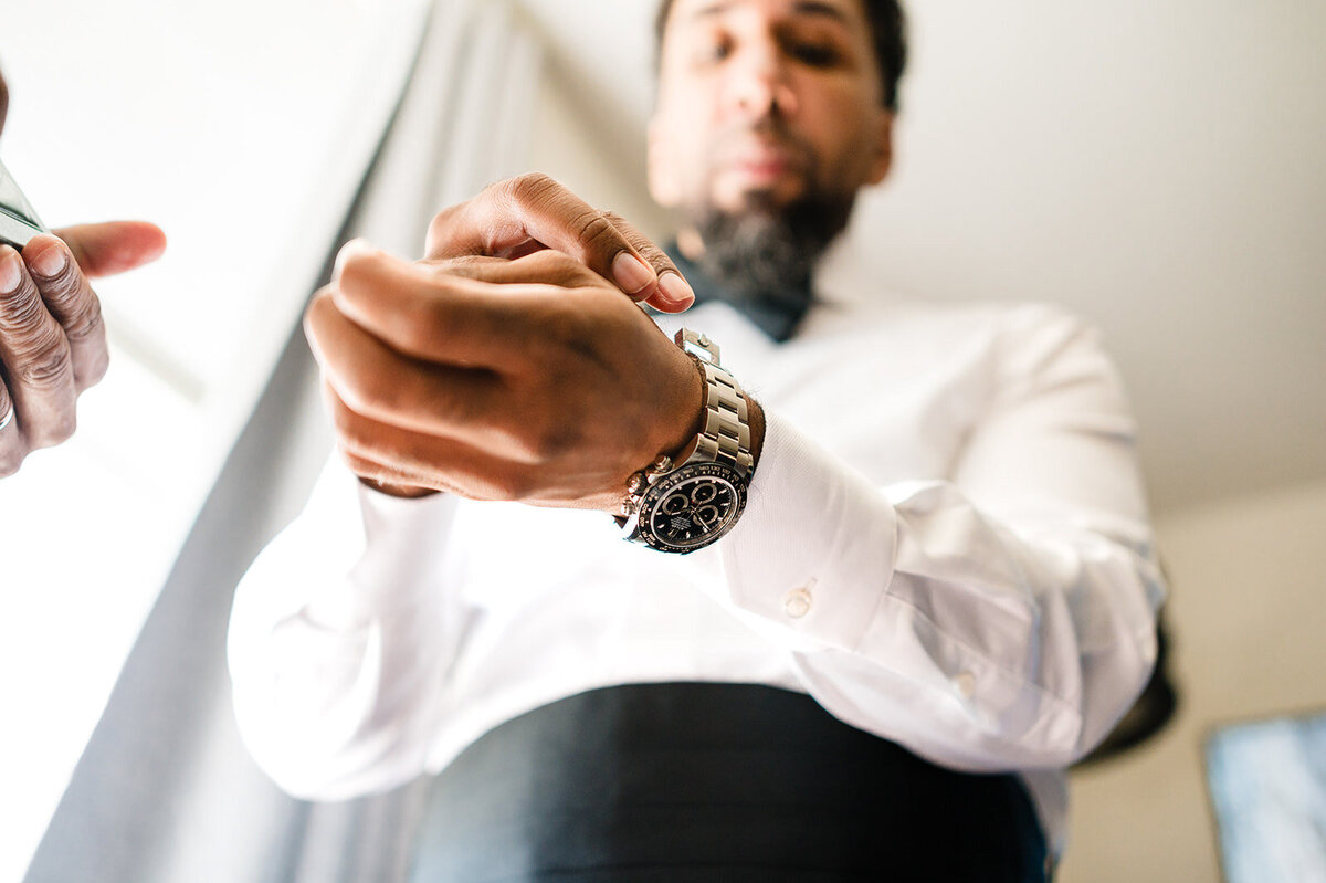 A groom in a white shirt, rolling up his sleeve to reveal a luxurious watch, standing by a window that casts a natural light on him